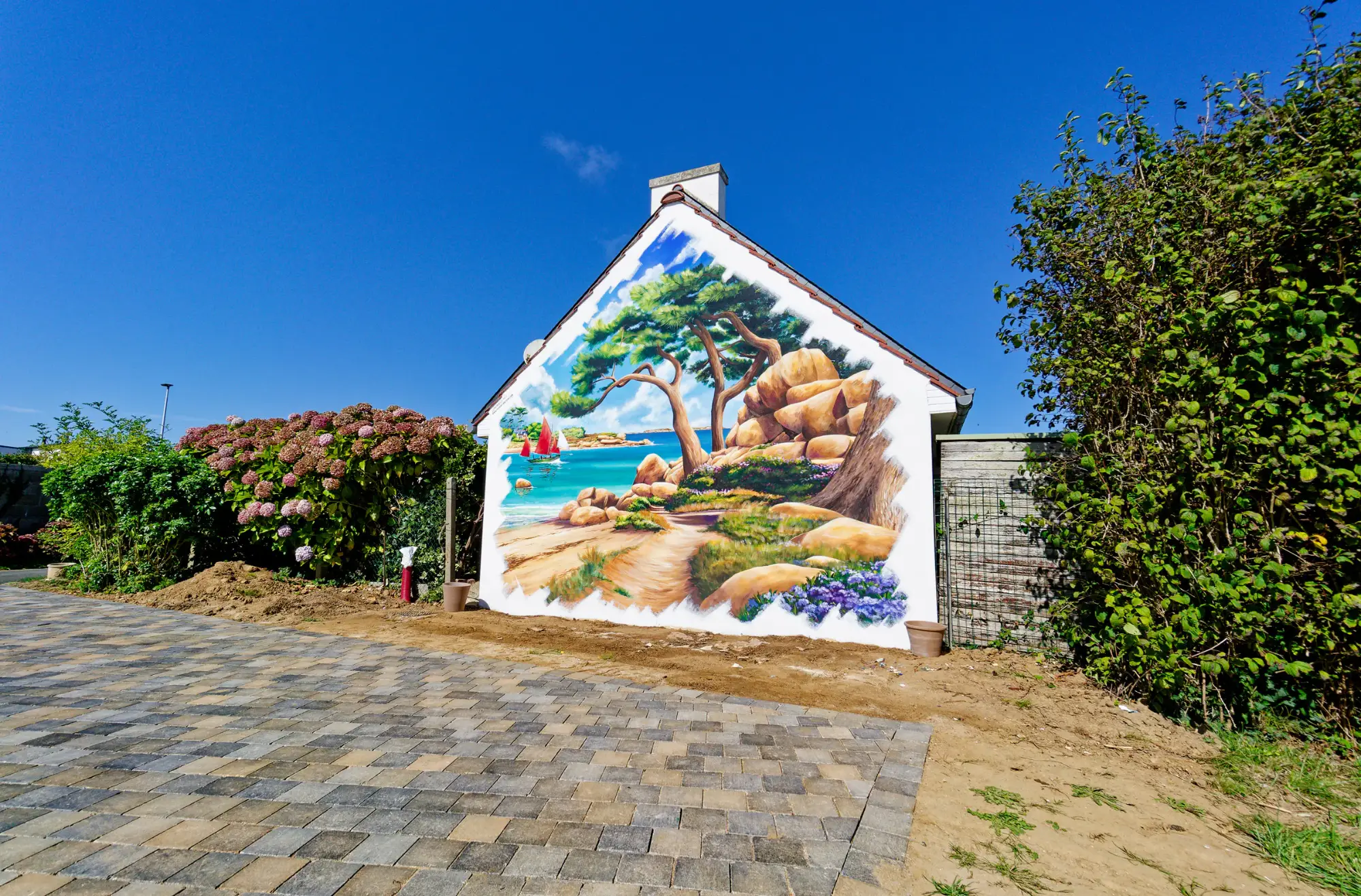 Vue large du pignon peint en trompe-l'œil, allée pavée et haie d'hortensias, mur mitoyen de jardin à Trégastel